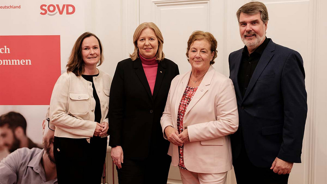 Vier Personen nebeneinander vor einem SoVD-Plakat. 