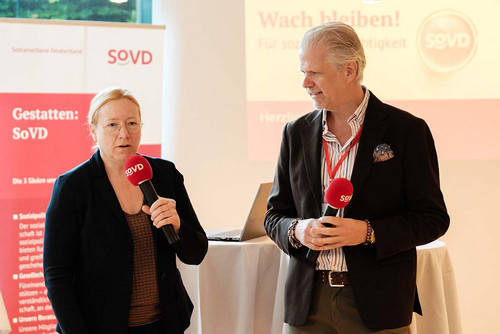 Dagmar Schmidt, Schirmherrin des parlamentarischen Frühstücks und SPD-Abgeordnete, spricht am Mikrofon. Neben ihr steht SoVD-Bundespressesprecher Peter-Michael Zernechel. Im Hintergrund ist an die Leinwand projiziert: "Wach bleiben! Für soziale Gerechtigkeit."