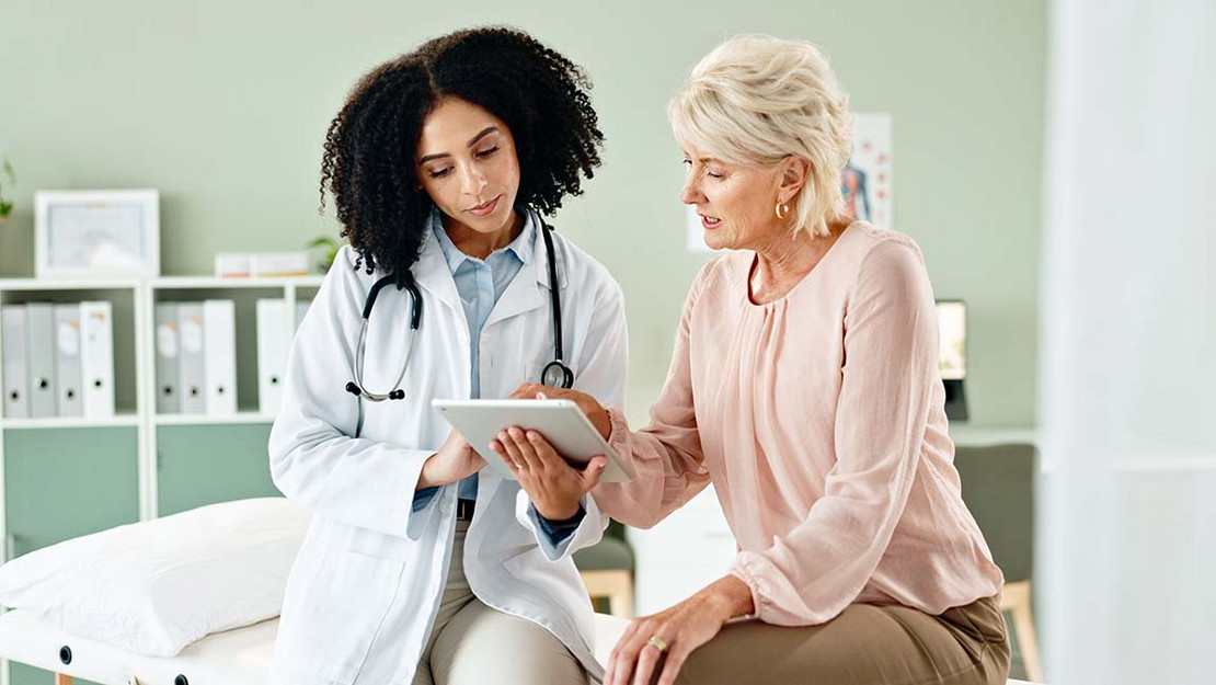 Eine Patientin sitzt neben einer Ärztin, die etwas auf dem Tablet zeigt. 