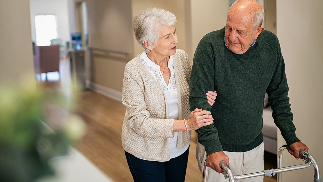 Ein älterer Mann mit Rollator und seine Frau laufen über den Gang eines Pflegeheims. 