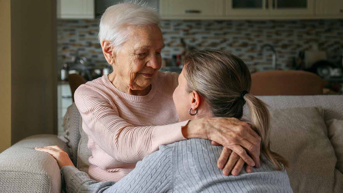 Eine ältere Frau sitzt auf dem Sofa und umschlingt den Hals einer jüngeren Frau. 