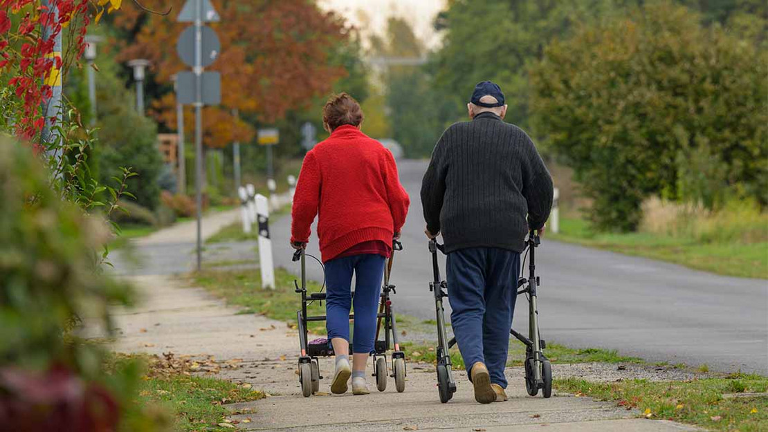 Rückenansicht: Rentnerpaar mit Rollatoren laufen auf einem Gehweh. 