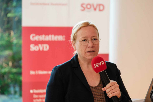 Frau steht vor einem SoVD-Aufsteller und spricht in ein Mikrofon.