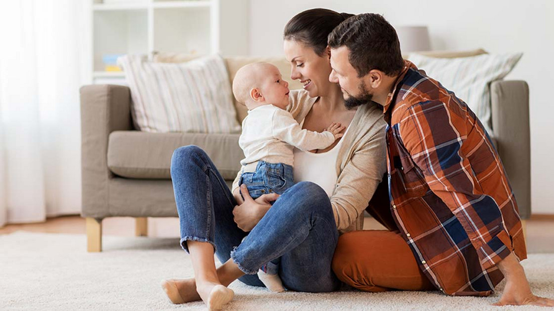 Junge Eltern sitzen in einer Wohnung auf dem Boden. Die Mutter hält eine Baby auf dem Arm. 
