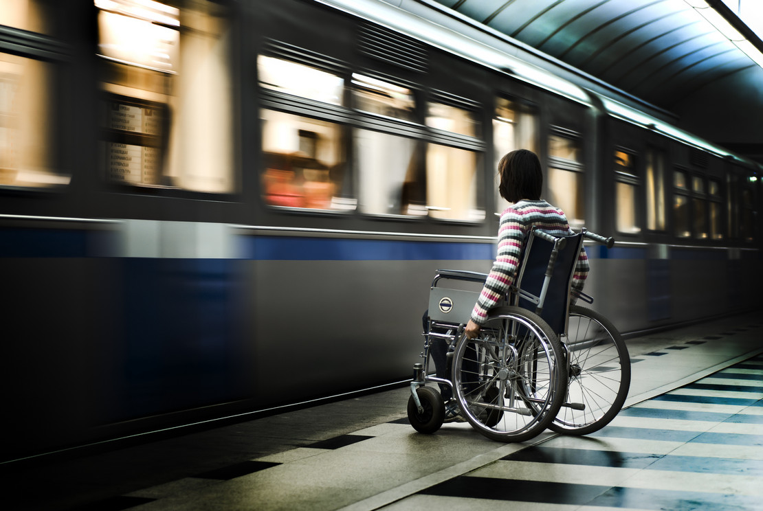 Frau im Rollstuhl vor einfahrendem Zug am Bahnsteig. 