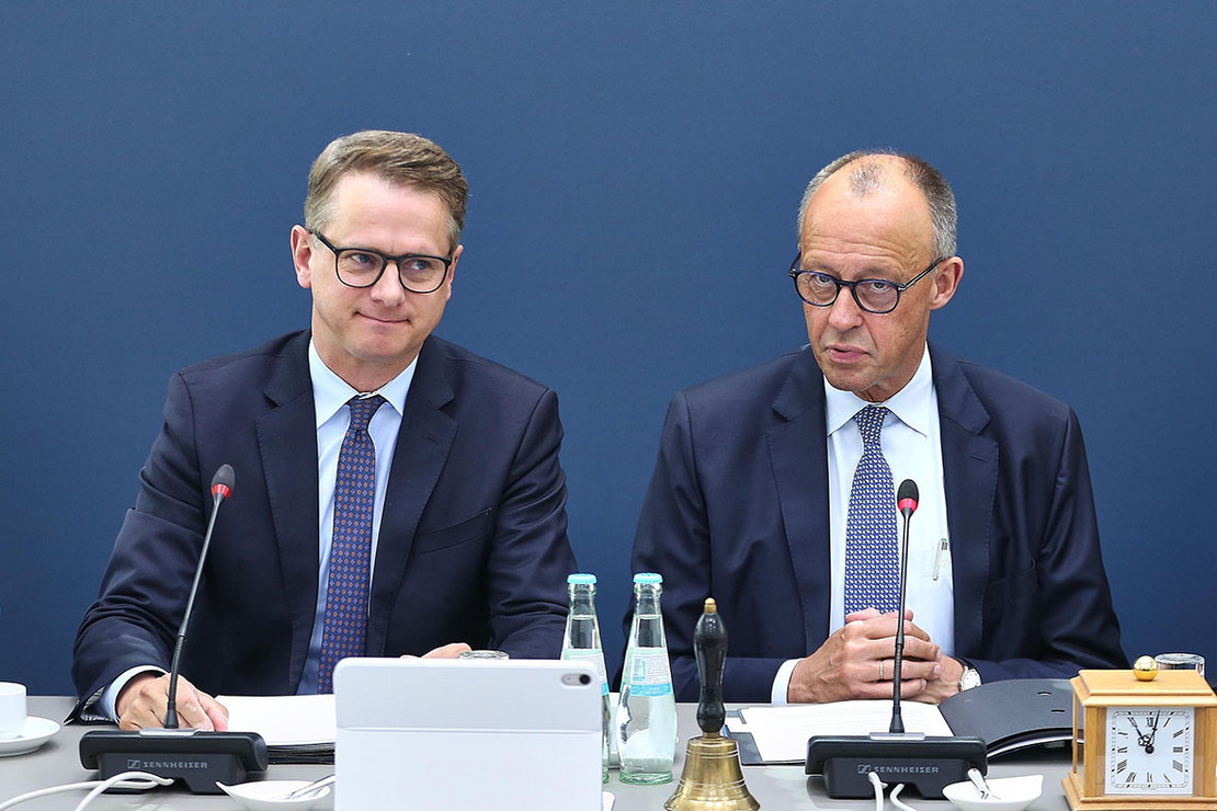 zwei Männer in Anzügen sitzen an einem Rednertisch