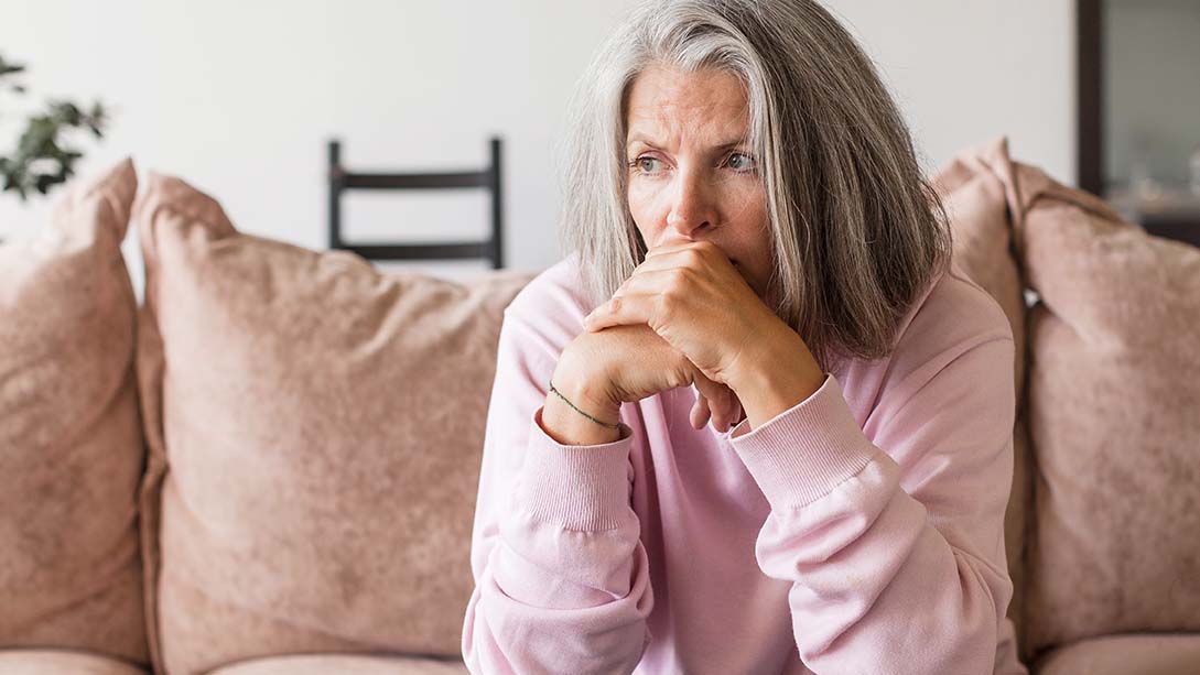 Mittelalte Frau sitzt auf einem Sofa und hat ein besorgtes Gesicht. 